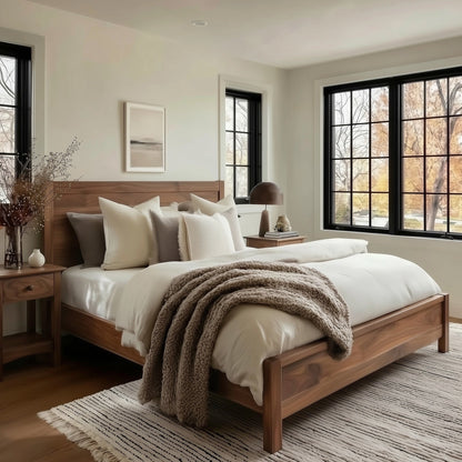 Neatly made bed with wooden headboard in a bedroom with large windows.